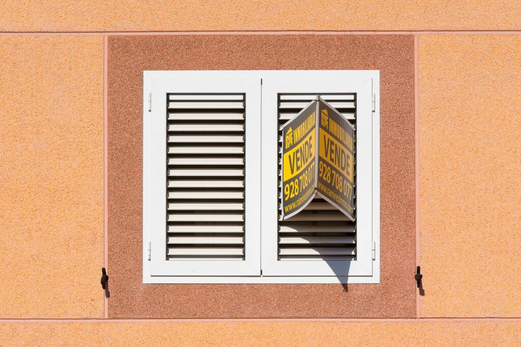 White shutter window with a for sale sign on peach building exterior.