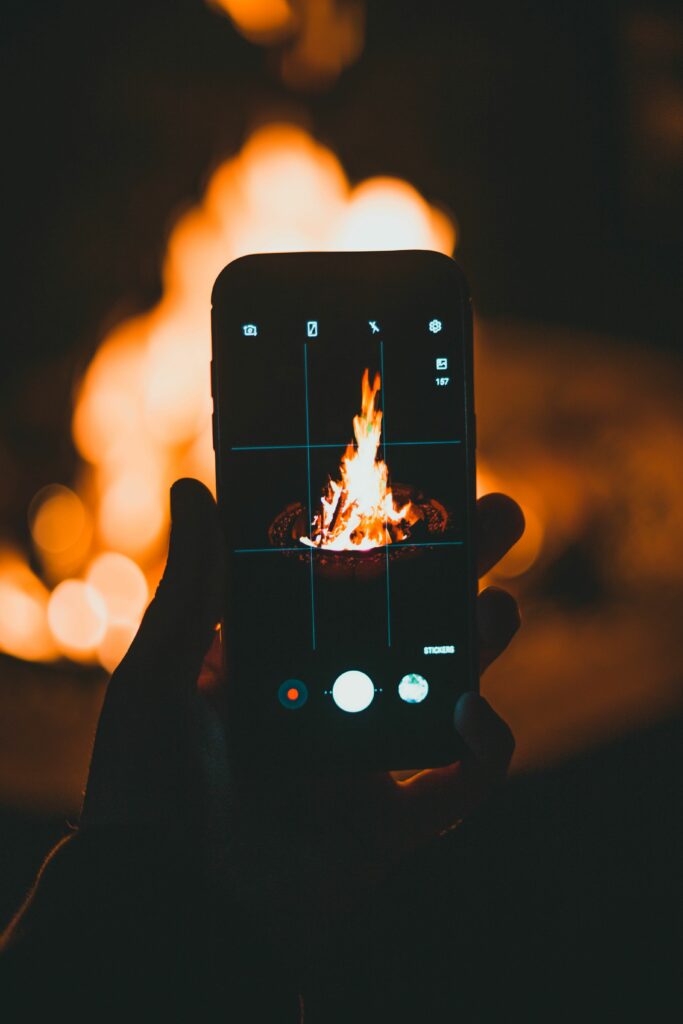 A hand holding a smartphone capturing a blazing campfire at night, creating a warm, cozy atmosphere.